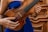 A person wearing a blue garment holds a brown wooden violin, with intricate woodwork in the background. The hands are positioned to hold and play the instrument, showcasing both the body and strings of the violin.
