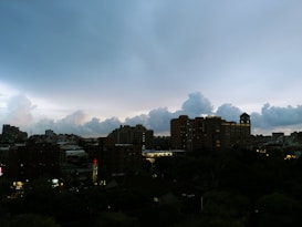 Dusk settles over an urban skyline, with a collection of mid-rise buildings silhouetted against a cloudy sky. Individuals glowing lights from buildings and a cross illuminated in red suggest various establishments. The horizon shows a mixture of clouds against a slowly darkening blue and purple sky.