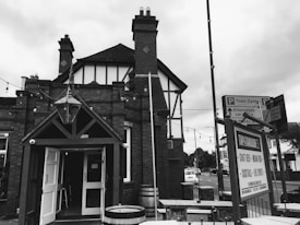 A black and white image of a traditional English-style pub or tavern with brick architecture and wooden accents. There are string lights hanging across the entrance and signage nearby directing to a town center and other locations. The atmosphere is quaint and slightly nostalgic.