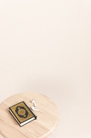A wooden round stool holding a book with an ornate black and gold cover and a pair of wireless earbuds. The background is a soft beige color.