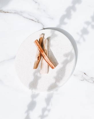Three pieces of wooden sticks are neatly arranged on a light-colored ceramic plate, set against a white marble surface. Shadows of leaves or branches are cast delicately on the setup, creating a serene and natural ambiance.