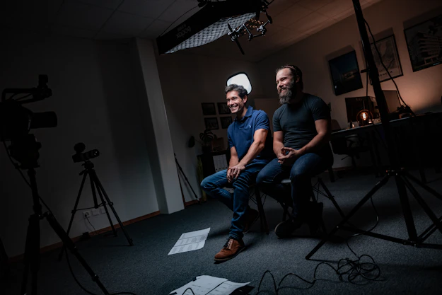 Two clothed male influencers collaborating over content planning, seated with laptops in a softly lit studio emphasizing professionalism.