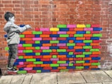 A brick wall features a colorful mural of various brightly painted bricks. To the left, a grayscale depiction of a child appears to be painting the bricks. The wall behind is made of traditional red bricks, with the painted section interrupting the uniformity with vibrant colors.
