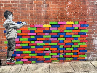 A brick wall features a colorful mural of various brightly painted bricks. To the left, a grayscale depiction of a child appears to be painting the bricks. The wall behind is made of traditional red bricks, with the painted section interrupting the uniformity with vibrant colors.