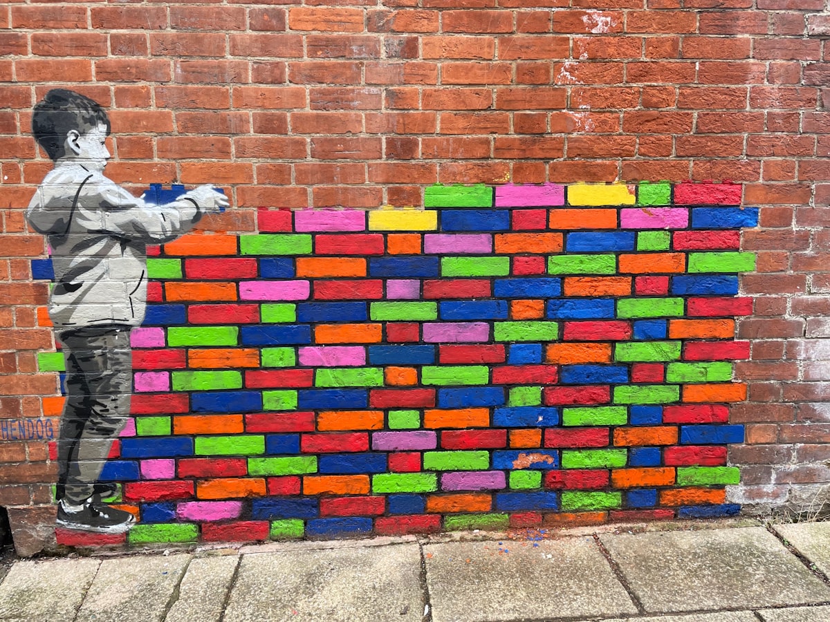 A red brick wall with a mural of a child in greyscale placing colorful blue, red, pink, yellow and green bricks in the wall