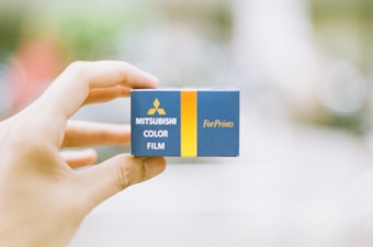 A hand holds a small blue and yellow box labeled with 'Mitsubishi Color Film ForPrints.' The background is blurred, focusing attention on the box.