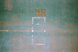A green textured wall with the words 'NO NUKE' stenciled in orange paint. The words are centered in the upper portion of the image, and there are faint outlines of shapes or patterns below them.