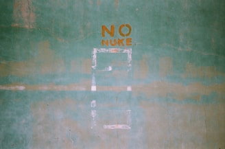 A green textured wall with the words 'NO NUKE' stenciled in orange paint. The words are centered in the upper portion of the image, and there are faint outlines of shapes or patterns below them.