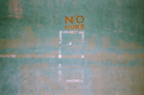 A green textured wall with the words 'NO NUKE' stenciled in orange paint. The words are centered in the upper portion of the image, and there are faint outlines of shapes or patterns below them.