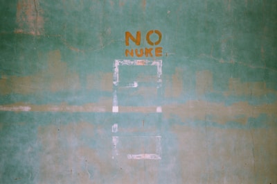 A green textured wall with the words 'NO NUKE' stenciled in orange paint. The words are centered in the upper portion of the image, and there are faint outlines of shapes or patterns below them.