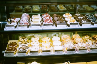 An assortment of beautifully decorated pastries and desserts displayed in a glass case. Each dessert is presented on individual trays with price tags, showcasing a variety of toppings such as berries, chocolate, and cream.