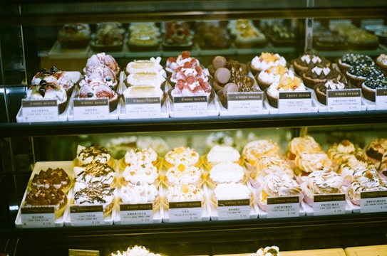 An assortment of beautifully decorated pastries and desserts displayed in a glass case. Each dessert is presented on individual trays with price tags, showcasing a variety of toppings such as berries, chocolate, and cream.