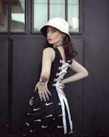 A person stands in front of a dark, industrial background, wearing a white bucket hat and a stylish black top with lace-up detailing on the side. The outfit is complemented by black pants featuring a pattern of white swoosh symbols, and accessorized with hoop earrings, a bracelet, and rings. The atmosphere is modern with a touch of street style.