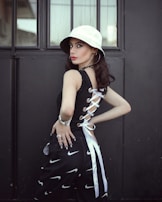 A person stands in front of a dark, industrial background, wearing a white bucket hat and a stylish black top with lace-up detailing on the side. The outfit is complemented by black pants featuring a pattern of white swoosh symbols, and accessorized with hoop earrings, a bracelet, and rings. The atmosphere is modern with a touch of street style.