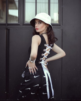 A person stands in front of a dark, industrial background, wearing a white bucket hat and a stylish black top with lace-up detailing on the side. The outfit is complemented by black pants featuring a pattern of white swoosh symbols, and accessorized with hoop earrings, a bracelet, and rings. The atmosphere is modern with a touch of street style.