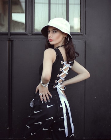 A person stands in front of a dark, industrial background, wearing a white bucket hat and a stylish black top with lace-up detailing on the side. The outfit is complemented by black pants featuring a pattern of white swoosh symbols, and accessorized with hoop earrings, a bracelet, and rings. The atmosphere is modern with a touch of street style.