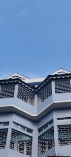 A three-story building with a modern architectural design featuring large windows and angled roof peaks. The facade is painted in a light blue color, and the building stands against a backdrop of clear blue sky.