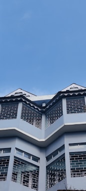 A three-story building with a modern architectural design featuring large windows and angled roof peaks. The facade is painted in a light blue color, and the building stands against a backdrop of clear blue sky.
