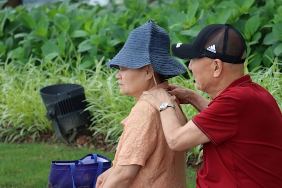 An older man is giving a shoulder massage to a woman who is wearing a blue sun hat. They are both seated outdoors on the grass, surrounded by lush green plants. The man is wearing a red polo shirt and a black cap, while the woman is in an orange textured blouse. Nearby, there is a blue bag with handles.