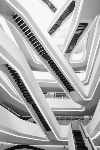 A modern architectural design featuring intersecting escalators forming geometric patterns. The structure is sleek with a monochrome palette, and the lines create a sense of movement and complexity.