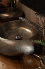 A metallic basin with a textured surface sits on a wooden countertop. The faucet is sleek and chrome-finished, complementing the basin's design. The setting is dimly lit, creating a reflective and slightly rustic ambiance.