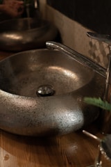A metallic basin with a textured surface sits on a wooden countertop. The faucet is sleek and chrome-finished, complementing the basin's design. The setting is dimly lit, creating a reflective and slightly rustic ambiance.