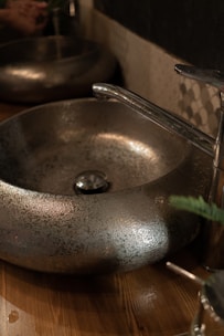 A metallic basin with a textured surface sits on a wooden countertop. The faucet is sleek and chrome-finished, complementing the basin's design. The setting is dimly lit, creating a reflective and slightly rustic ambiance.