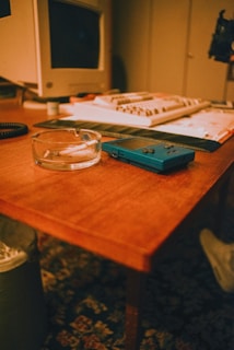 A vintage setup featuring a wooden table with a transparent ashtray containing a cigarette, an old handheld gaming console, and a cluttered desktop with a tube monitor and keyboard in the background. The atmosphere suggests a retro or nostalgic setting.