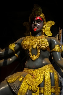 A statue of a deity with a dark complexion, adorned with intricate golden jewelry, including ornate earrings, necklaces, and a headpiece. The figure's eyes are accentuated with red and the tongue is sticking out, adding to the intense expression.