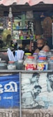 A small shop is filled with various items, including snacks and household products, neatly stacked on shelves. A man wearing a face mask is sitting inside the shop. The counter in the foreground displays jars containing snacks and plastic containers. A fan and light bulb are visible, along with a poster featuring people using mobile phones.