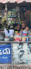 A small shop is filled with various items, including snacks and household products, neatly stacked on shelves. A man wearing a face mask is sitting inside the shop. The counter in the foreground displays jars containing snacks and plastic containers. A fan and light bulb are visible, along with a poster featuring people using mobile phones.