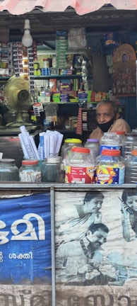 A small shop is filled with various items, including snacks and household products, neatly stacked on shelves. A man wearing a face mask is sitting inside the shop. The counter in the foreground displays jars containing snacks and plastic containers. A fan and light bulb are visible, along with a poster featuring people using mobile phones.