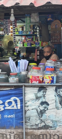 A small shop is filled with various items, including snacks and household products, neatly stacked on shelves. A man wearing a face mask is sitting inside the shop. The counter in the foreground displays jars containing snacks and plastic containers. A fan and light bulb are visible, along with a poster featuring people using mobile phones.