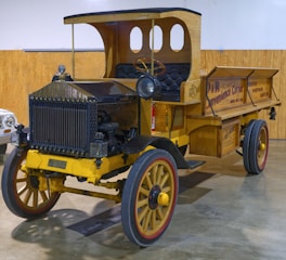 A vintage wooden delivery truck with large wooden wheels and a yellow chassis is displayed in an indoor setting. The cab and cargo area feature intricate woodwork, and the truck has an old-fashioned headlight on the front. The side of the truck bed displays vintage advertising for a convenience corner and groceries, adding to its historical charm.