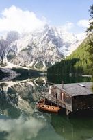 A serene mountain landscape with towering snow-capped peaks reflected in a clear, still lake. A rustic wooden cabin on stilts sits by the water's edge, with wooden boats moored nearby. Lush green forests line the shore, and fluffy white clouds drift overhead.