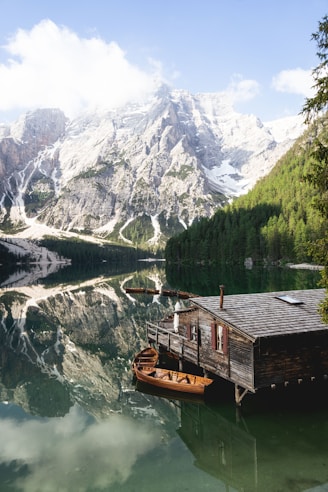A serene mountain landscape with towering snow-capped peaks reflected in a clear, still lake. A rustic wooden cabin on stilts sits by the water's edge, with wooden boats moored nearby. Lush green forests line the shore, and fluffy white clouds drift overhead.