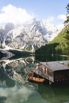 A serene mountain landscape with towering snow-capped peaks reflected in a clear, still lake. A rustic wooden cabin on stilts sits by the water's edge, with wooden boats moored nearby. Lush green forests line the shore, and fluffy white clouds drift overhead.