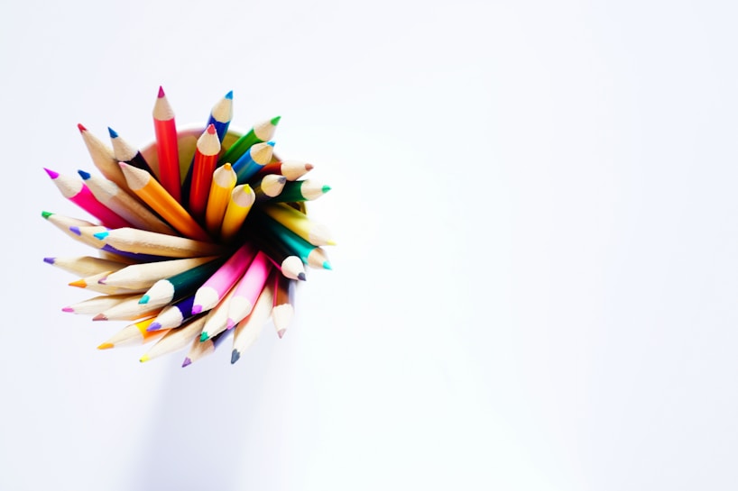 A collection of colored pencils arranged in a circular pattern, with the sharpened tips pointing upward. The pencils display a variety of vibrant colors against a plain white background, creating a striking contrast.