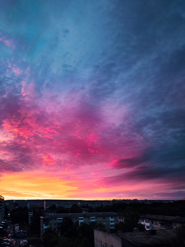 The image captures a vibrant and colorful sunset over a cityscape, with dramatic clouds displaying a mix of pink, purple, and orange hues. The buildings below are shadowed, creating a stark contrast against the illuminated sky.