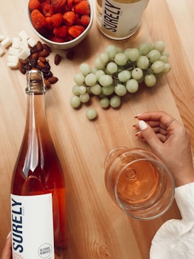 A wooden table displays an assortment of food and drink items, including a bowl of raspberries, cubes of white cheese, almonds, a bunch of green grapes, and a bottle of non-alcoholic rosé labeled 'Surely' with a corresponding glass of the drink nearby. A hand with manicured nails elegantly holds the glass, enhancing the serene and luxurious ambiance.