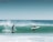 Two surfers are riding a wave; one is standing on the board while the other is lying down, preparing to catch the wave. The ocean water is a vibrant shade of turquoise, and the sky is a clear blue, creating a serene background. The waves are crashing with white foam, indicating a lively sea.