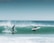 Two surfers are riding a wave; one is standing on the board while the other is lying down, preparing to catch the wave. The ocean water is a vibrant shade of turquoise, and the sky is a clear blue, creating a serene background. The waves are crashing with white foam, indicating a lively sea.