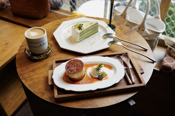 A wooden table is set with a cup of cappuccino, a slice of layered green tea cake with utensils, and a piece of caramel flan with whipped cream and a small piece of fruit garnish on a white plate. The items are elegantly arranged, creating a cozy and inviting café atmosphere.