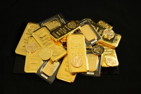 A pile of gold bars and coins scattered on a dark surface, featuring inscriptions and logos, some bars are stacked neatly while others are overlapping.