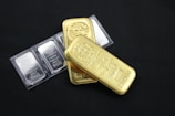Several gold and platinum bars are arranged on a dark background. The gold bars have markings indicating 500g fine gold, while the platinum bars are marked as 1 ounce of fine platinum. The shiny metallic surfaces reflect light.