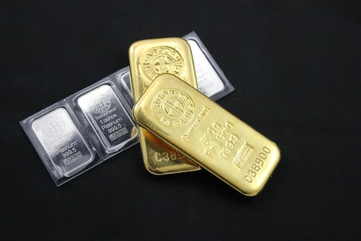 Several gold and platinum bars are arranged on a dark background. The gold bars have markings indicating 500g fine gold, while the platinum bars are marked as 1 ounce of fine platinum. The shiny metallic surfaces reflect light.