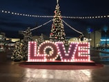 Large illuminated letters spell out the word 'LOVE' in front of a decorated Christmas tree at an outdoor plaza. String lights create a festive atmosphere, with additional smaller trees enhancing the holiday spirit. The scene takes place during the evening, with lights brightening the surroundings.