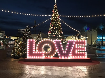 Large illuminated letters spell out the word 'LOVE' in front of a decorated Christmas tree at an outdoor plaza. String lights create a festive atmosphere, with additional smaller trees enhancing the holiday spirit. The scene takes place during the evening, with lights brightening the surroundings.