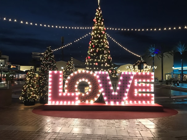 Large illuminated letters spell out the word 'LOVE' in front of a decorated Christmas tree at an outdoor plaza. String lights create a festive atmosphere, with additional smaller trees enhancing the holiday spirit. The scene takes place during the evening, with lights brightening the surroundings.
