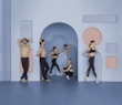 A group of dancers is positioned in a minimalistic setting with geometric shapes on the walls. The dancers are dressed in black dance apparel, with some performing ballet poses and others resting. The setting features a color scheme of blue and pink.